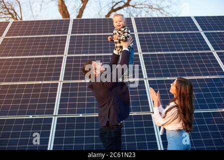 Un jeune homme jette un petit enfant sur un fond de panneaux solaires sous un ciel bleu. Une belle femme aux cheveux longs est debout à proximité. Homme dans une veste. Image de concept d'énergie solaire Banque D'Images