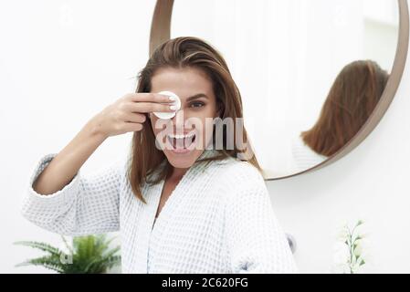 Une jeune fille gaie avec un sourire blanc neige tient un tampon de coton près de ses yeux et rit. Une jolie brunette nettoie sa peau avec un tampon de coton avec de l'eau micellaire ou un nettoyant. Banque D'Images