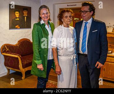 Trois générations de la famille d’hôteliers Metzler se tiennent à l’entrée du restaurant du Romantik Hotel Das Schiff à Hittisau, Vorarlberg, Autriche : Antonia Metzler (g.) et son mari Hans-Georg Metzler (d.), le couple propriétaire actuel, encadrent sa mère Erna Metzler (au centre) dans ses vêtements de cuisine blancs. Un portrait des grands-parents est accroché au mur derrière eux. Banque D'Images