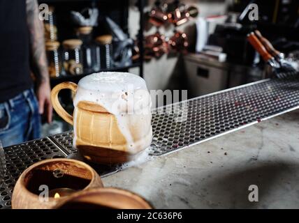 Bière artisanale froide dans une tasse en céramique avec une goutte de mousse. Un barman mâle debout près du comptoir du bar dans le pub Banque D'Images