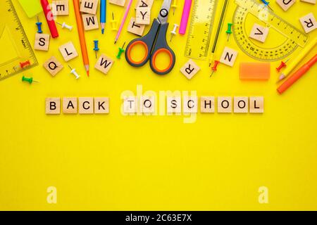 Lettres en bois disposées en phrase de retour à l'école, crayons, stylos-feutres, épingles, ciseaux et fournitures de bureau sur fond jaune. Banque D'Images