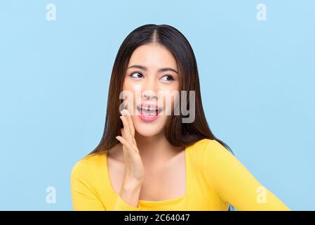 Jeune femme asiatique qui dit quelque chose avec la main à côté de la bouche sur fond bleu clair isolé Banque D'Images