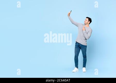 Portrait complet d'un jeune homme beau qui prend le selfie sur un téléphone portable isolé sur un fond bleu clair avec un espace de copie Banque D'Images