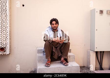 Nizwa / Oman - 15 février 2020 : homme immigrant musulman assis au téléphone dans la rue de Nizwa Banque D'Images