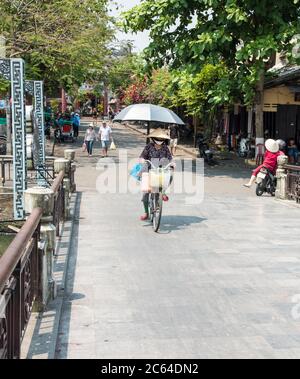 Femme vietnamienne locale portant un masque facial et faisant du vélo sur un pont dans la ville ancienne de Hoi an Vietnam Asie. Banque D'Images