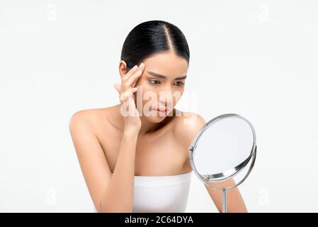 Jeune femme asiatique belle regardant dans le miroir et touchant son visage pour vérifier les rides isolées sur fond blanc pour les soins de la peau et de la beauté concep Banque D'Images