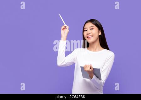 Souriante belle femme asiatique en tenue blanche pointant avec un stylet pour copier l'espace et tenant la tablette numérique isolée sur fond violet Banque D'Images