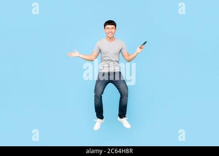 Portrait amusant en longueur d'un homme asiatique souriant qui saute en plein air tout en tenant son smartphone dans un arrière-plan bleu studio isolé Banque D'Images