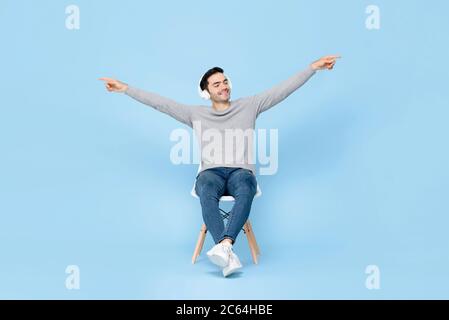 Portrait de la jeune femme de race blanche, reposant, assis sur une chaise, portant un casque d'écoute, écoutant de la musique avec les bras levés dans un arrière bleu de studio isolé Banque D'Images