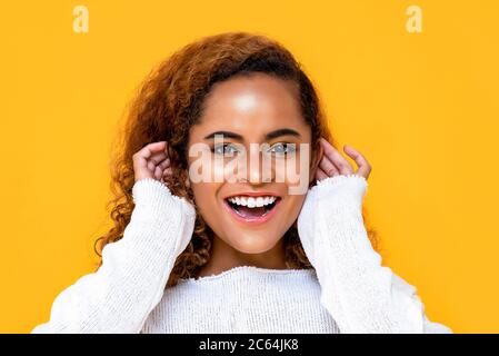 Portrait en gros plan d'une jeune femme afro-américaine joyeuse souriant tout en touchant ses oreilles des deux mains sur fond jaune studio isolé Banque D'Images