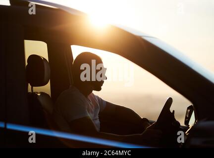 Silhouette d'un jeune homme assis dans sa voiture, portant des lunettes de soleil et regardant le coucher du soleil, main sur la barre, soleil brillant sur le toit de la voiture, gros plan, vue latérale Banque D'Images