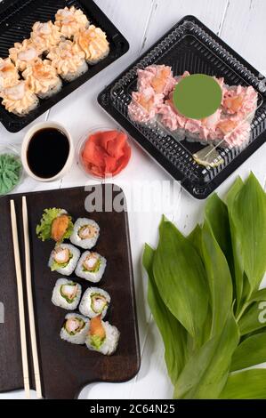 Des petits pains à sushis sont servis sur un panneau en bois sombre et des boîtes en plastique. Livraison de nourriture dans le restaurant dans des boîtes à emporter. Fond gris avec feuilles vertes Banque D'Images