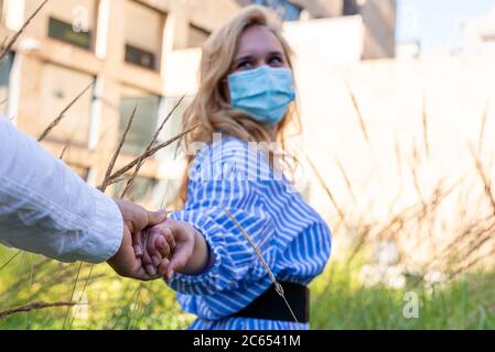 une jeune femme portant le masque protecteur, prend son homme à la main, l'invite à marcher vers un espace plus lumineux à travers les plantes, un im conceptuel Banque D'Images