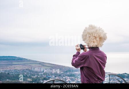 Touriste dans le noir rouge-rouge chemise et dans le traditionnel chapeau caucasien en peau de mouton blanc prend une photo du paysage de la mer et de la ville de Gagra, ABK Banque D'Images