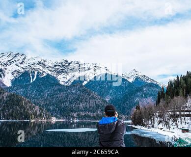 Un homme dans une veste noire avec une capuche bleue prend une photo d'un magnifique paysage d'hiver des montagnes du Caucase et du lac Ritsa, situé en Abkhazie Banque D'Images