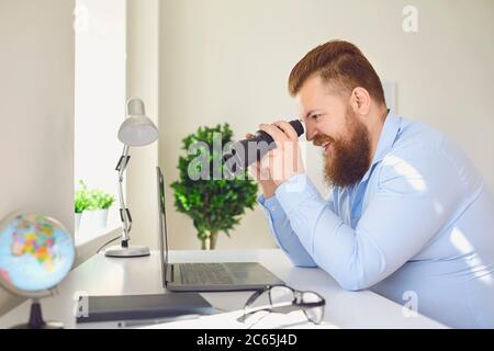 Drôle homme à la recherche de jumelles un ordinateur portable sur la table de travail à la maison bureau. Banque D'Images