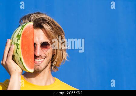 Jeune homme attrayant avec de longs cheveux blonds avec une moitié de pastèque couvrant son visage sur un fond bleu clair. Été, soleil, chaleur, fruits, conc. Vacances Banque D'Images