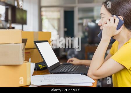 Femme d'affaires de PME qui parle sur téléphone portable avec ordinateur portable ouvert et colis sur table. Banque D'Images