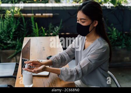 Vue latérale de la jeune femme asiatique sous masque de protection en appliquant de l'assainisseur sur les mains tout en étant assise à table sur la terrasse pendant une pandémie Banque D'Images