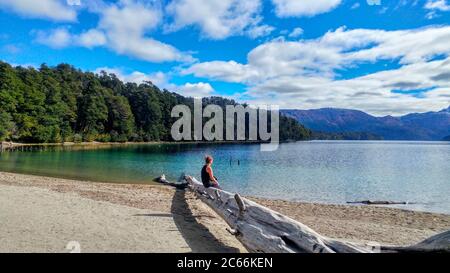 Femme assise sur un tronc d'arbre mort au bord du lac, face aux montagnes à l'horizon, Argentine Banque D'Images