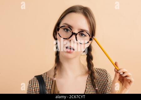 Portrait d'une jeune fille avec deux tresses dans des lunettes modernes tenant un crayon près de la tête regardant avec attention dans l'appareil photo sur fond beige Banque D'Images