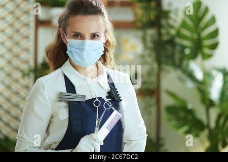 Survie des petites entreprises après une pandémie de Covid-19. Portrait de la femme propriétaire de petite entreprise élégante en tablier avec masque médical, gants, peigne à cheveux et scas Banque D'Images