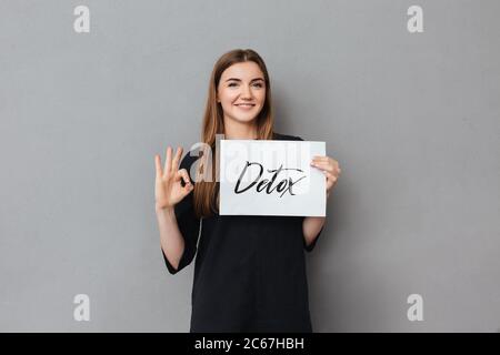 Portrait d'une jolie dame souriante debout et tenant une carte postale tout en regardant avec plaisir dans l'appareil photo sur fond gris Banque D'Images