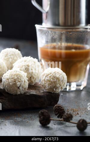 Boules d'énergie sucrées avec des flocons de noix de coco en pile sur pierre sombre avec du café phin vietnamien sur fond noir avec des petits cônes Banque D'Images