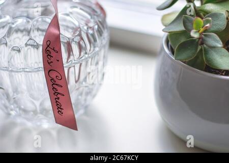 Pot décoratif en verre rétro avec un motif avec un ruban rose avec texte célébrons à côté d'une maison succulente sur un rebord de fenêtre blanc. Banque D'Images