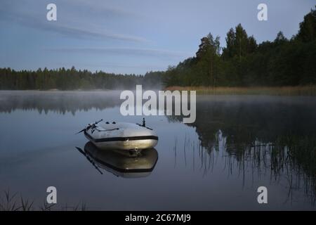 Le brouillard se curling sur les rives gazées du lac à l'aube, un bateau gonflable est en premier plan, région de Tver, Russie Banque D'Images