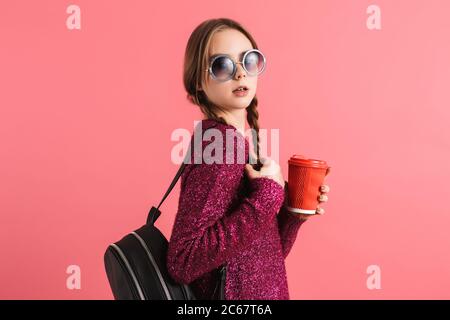 Jeune fille belle avec deux tresses dans le chandail et lunettes de soleil avec sac à dos noir tenant une tasse de café à aller à la main regardant soigneusement dans l'appareil photo Banque D'Images