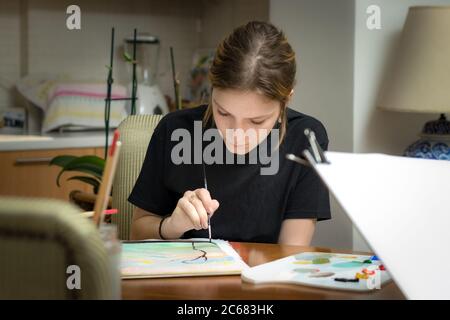 Une adolescente peint sur papier avec de l'acrylique tenant une brosse à peinture assise à la maison Banque D'Images