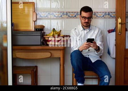 Un jeune homme regarde son téléphone assis dans une cuisine chaise Banque D'Images