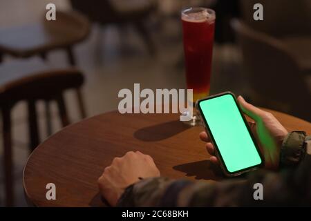 Sur l'épaule d'un homme tenant un smartphone à écran vert dans le bar la nuit. Un verre de cocktail sur une table de basse. Arrière-plan flou Banque D'Images