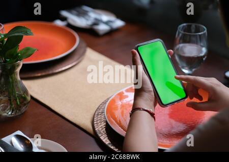 sur l'épaule des personnes qui tapotant un smartphone à écran vert dans le restaurant. assiettes floues et verre à boire sur la table. Banque D'Images