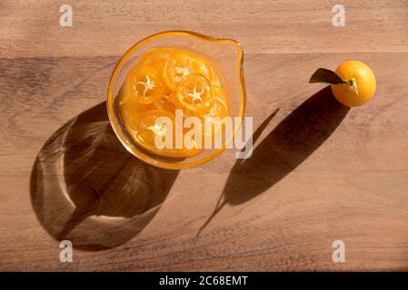 Un kumquat frais et d'autres confits et tranchés dans une petite soucoupe de verre, sur une surface en bois. Les deux émettent des ombres allongées et frappantes. Vue de dessus. Banque D'Images