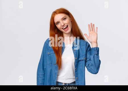 Sympathique, gaie et sortant jolie fille à tête rouge waving Palm, dire bonjour, saluant membre de l'équipe, incliner la tête et sourire joyeusement, gentil vous rencontrer Banque D'Images