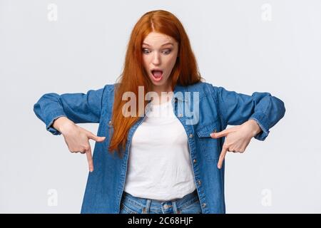 Portrait à la taille haute fille gaie impressionnée, curieuse et étonnante à tête rouge en denim, à mâchoire basse, regardant et pointant vers le bas étoilé sans voix Banque D'Images