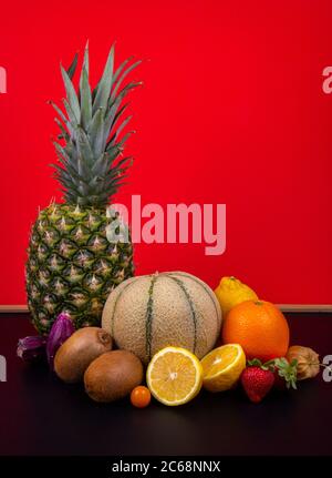 Un melon de miel, un ananas, et l'orange, des citrons, des kiwis, des figues de Barbarie et des lanternes chinoises devant un mur rouge. Banque D'Images