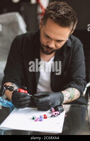 Tatoueur professionnel en gants noirs préparant la machine de tatouage pour le travail avec des encres colorées en studio Banque D'Images