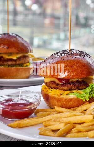 Deux hamburgers avec côtelettes de bœuf, fromage, frites, tomate, salade, sauce et ketchup sur une assiette blanche et un fond en bois clair Banque D'Images