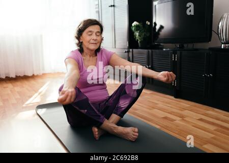 Femme sénior qui fait du yoga à la maison. femme de 70 ans assise sur un tapis de fitness avec les mains étirées sur ses genoux, effectuant des séances de yoga à liv Banque D'Images