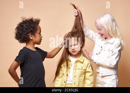 portrait de garçon à peau sombre et de fille albino touchant les cheveux longs d'ami caucasien, les jeunes coiffeurs. les enfants beaux avec l'apparence inhabituelle iso Banque D'Images