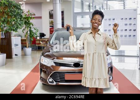 souriante femme africaine avec les clés de sa nouvelle voiture, elle est heureuse dans la concession, voiture de luxe dans l'arrière-plan Banque D'Images