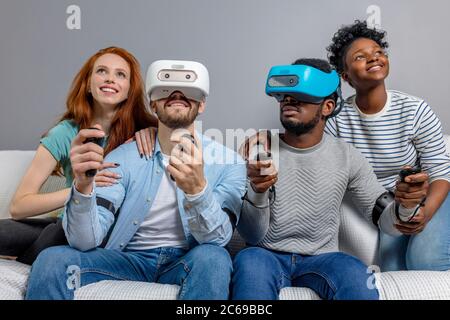 Deux hommes de différentes origines raciales ethnicité portant casque VR, jouant en studio de jeu et leurs copines les soutient, assise à côté de la table et l'chee Banque D'Images