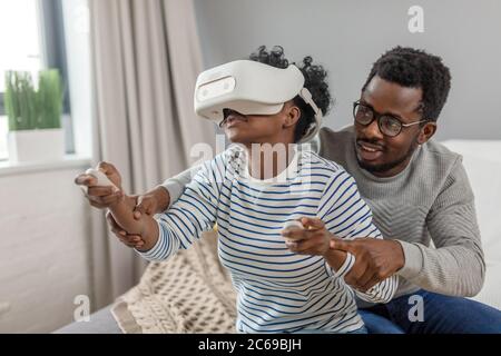 Beau couple à l'aide de dispositifs électroniques de technologie futuriste. Femme africaine jouant jeu d'ordinateur à couper le souffle avec casque VR et son mari il Banque D'Images