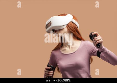 Gingembre occasionnels-haired woman en violet pullower portant casque VR et à impliqués dans le jeu de simulation 3D, désignant les mains comme s'il en interaction avec Banque D'Images