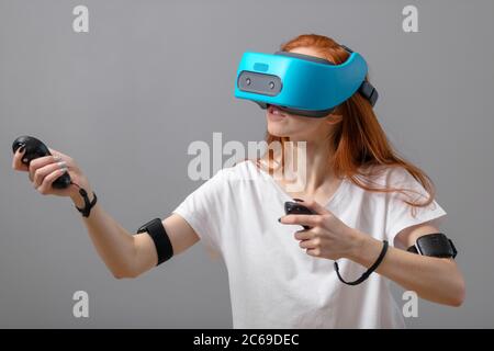 Femme rousse en t-shirt blanc portant l'aide du casque de réalité virtuelle, l'expérience tout en jouant des jeux vidéo, sur fond gris isolé Banque D'Images