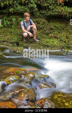 Un jeune homme (MR) croque à côté d'un petit ruisseau traversant une forêt dans le nord de Maui, Hawaï. Banque D'Images