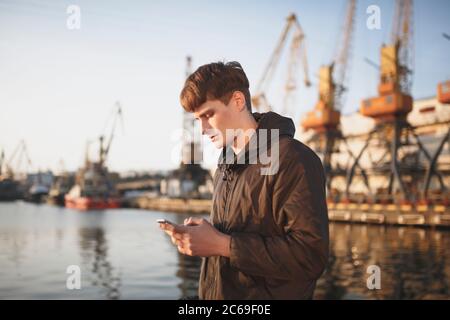 Jeune homme aux cheveux bruns debout et regardant avec soin dans son téléphone portable. Cool garçon dans la veste en duvet debout avec le téléphone cellulaire dans les mains avec la mer Banque D'Images